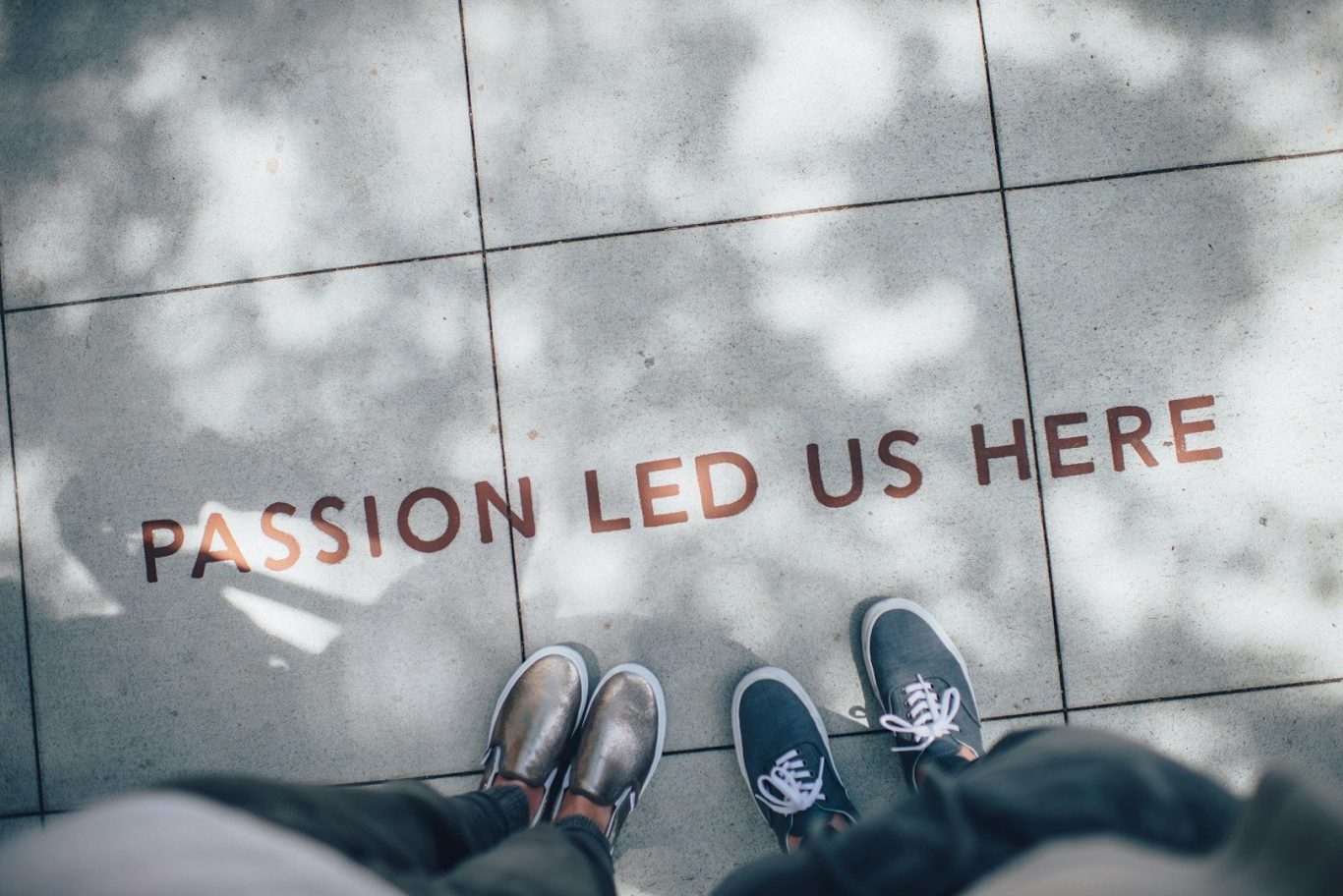 Two feet standing in front of chalk written on sidewalk that says, "Passion Led us Here"